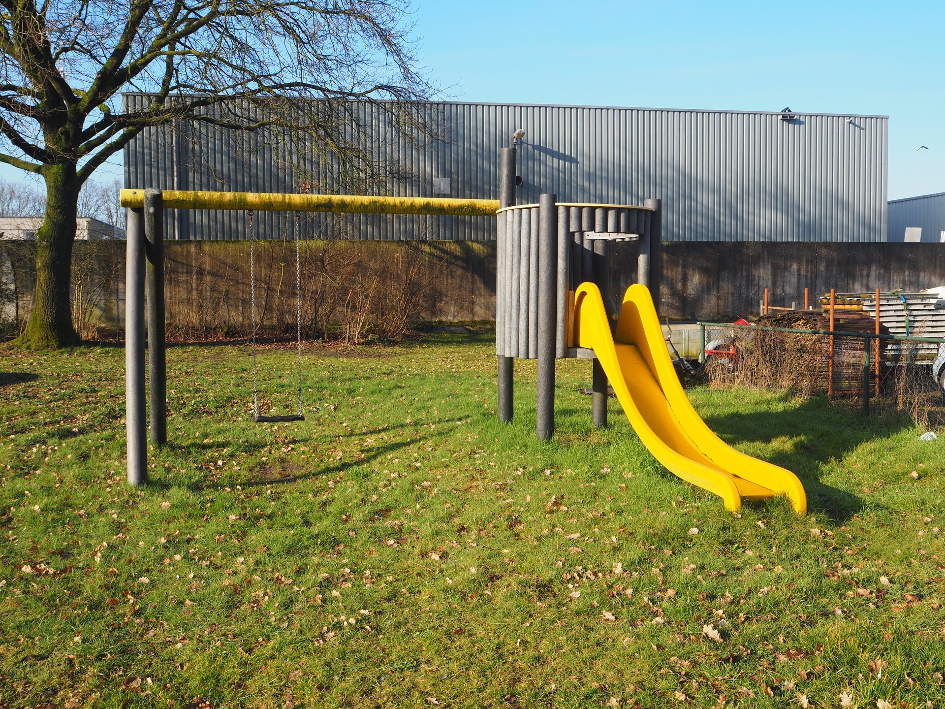 Vergroot afbeelding: aan de linkerkant een schommel met rechts daarvan een glijbaan, op de achtergrond staat een bedrijfsgebouw