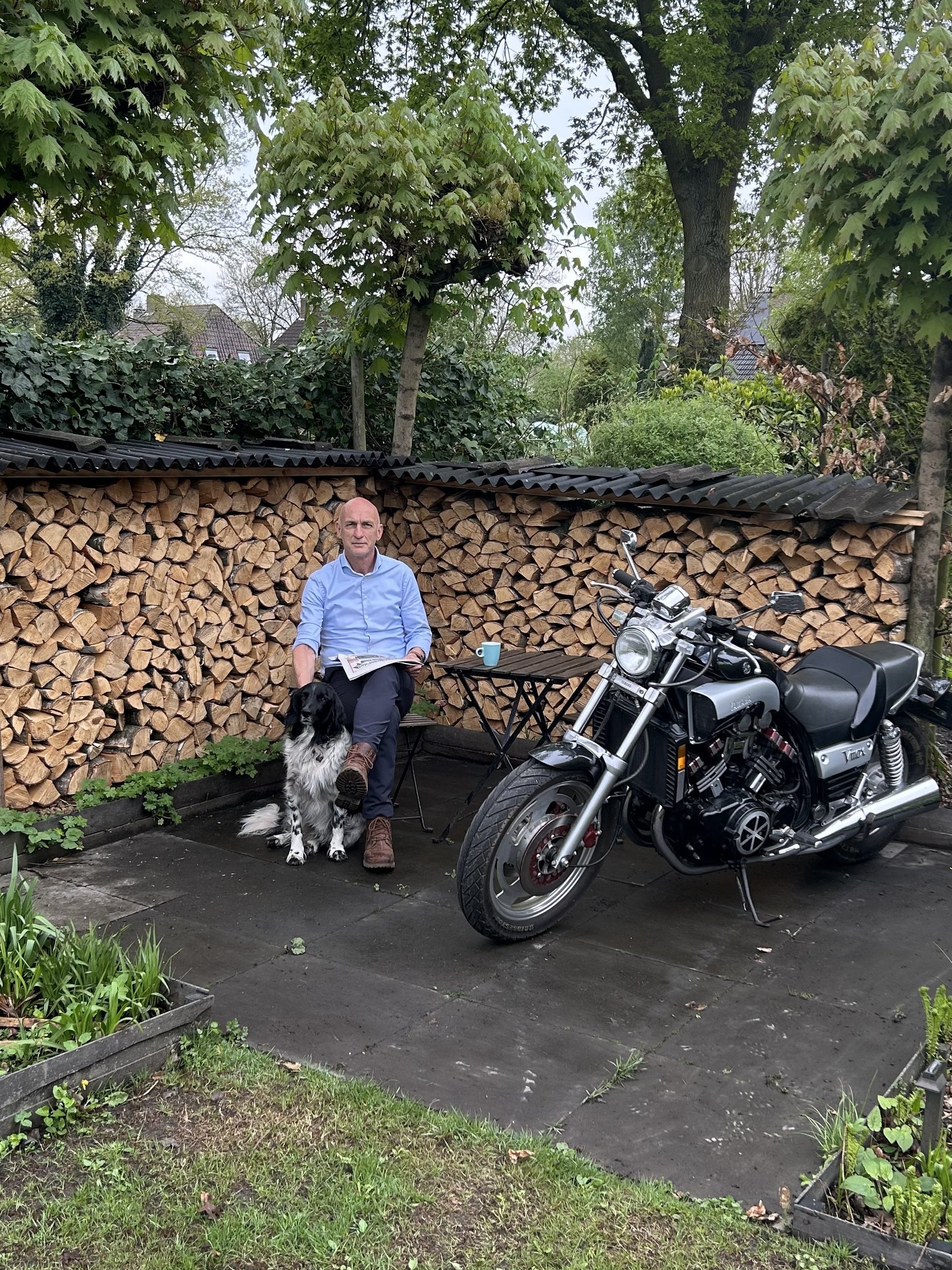 Vergroot afbeelding: gemeenteraadslid Willie van den Heuvel met voor hem zijn hond. Aan de rechterkant staat zijn motor en op de achtergrond is haardhout te zien.