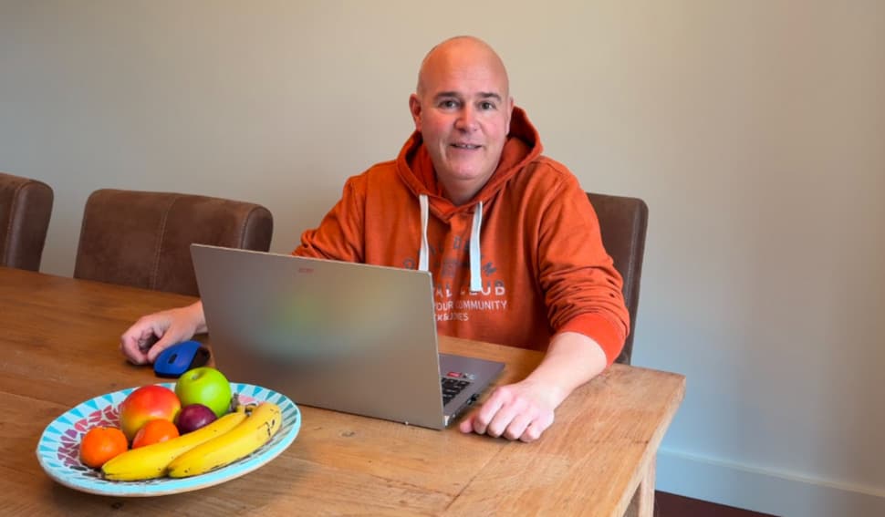 Vergroot afbeelding: cursist Fred in een oranje sweater achter zijn laptop. Linksonder staat een blauwe fruitmand met daarin fruit.